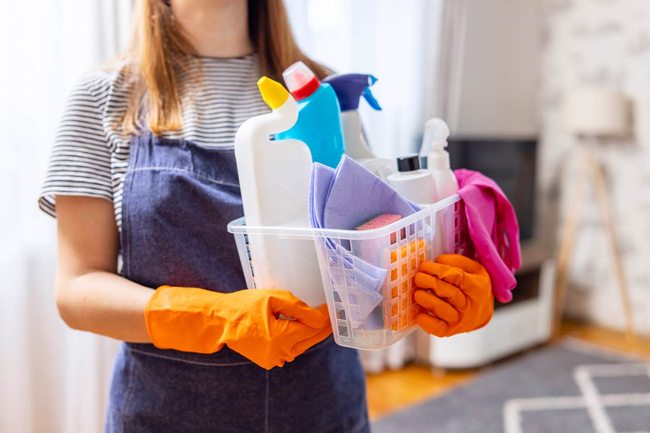 woman in rubber gloves with basket of cleaning supplies ready to clean up her apartment